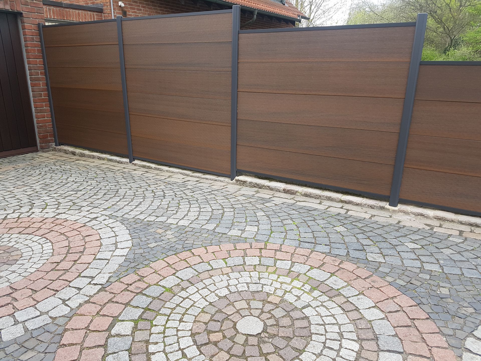 Brauner Sichtschutzzaun mit dunkelgrauen Rahmen auf einer gepflasterten Auffahrt.
