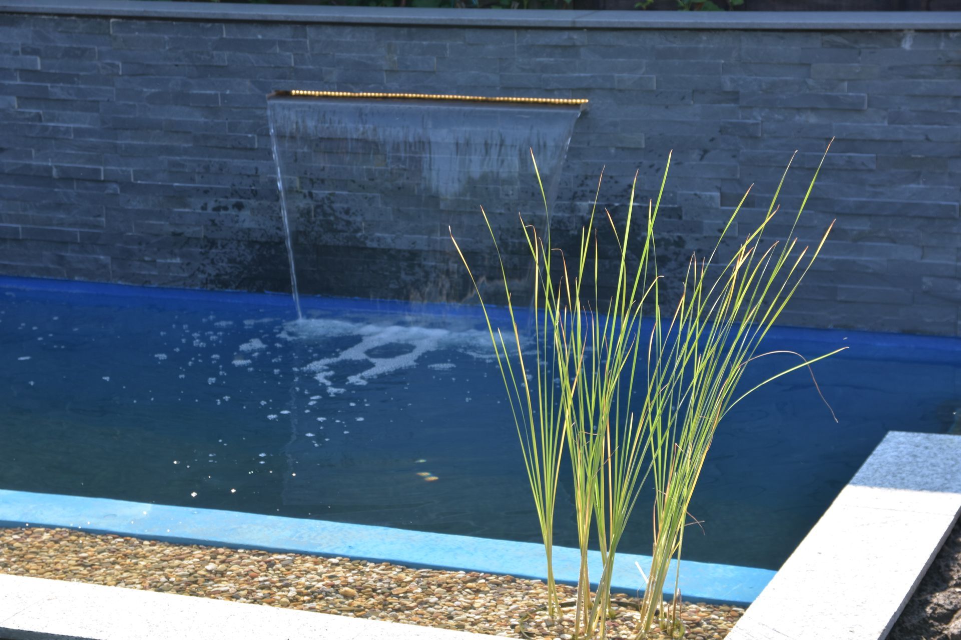 Wasserspiel mit Wasserfall, der in ein blau umrandetes Becken fließt. Hohes Gras im Vordergrund, Steinmauer im Hintergrund.