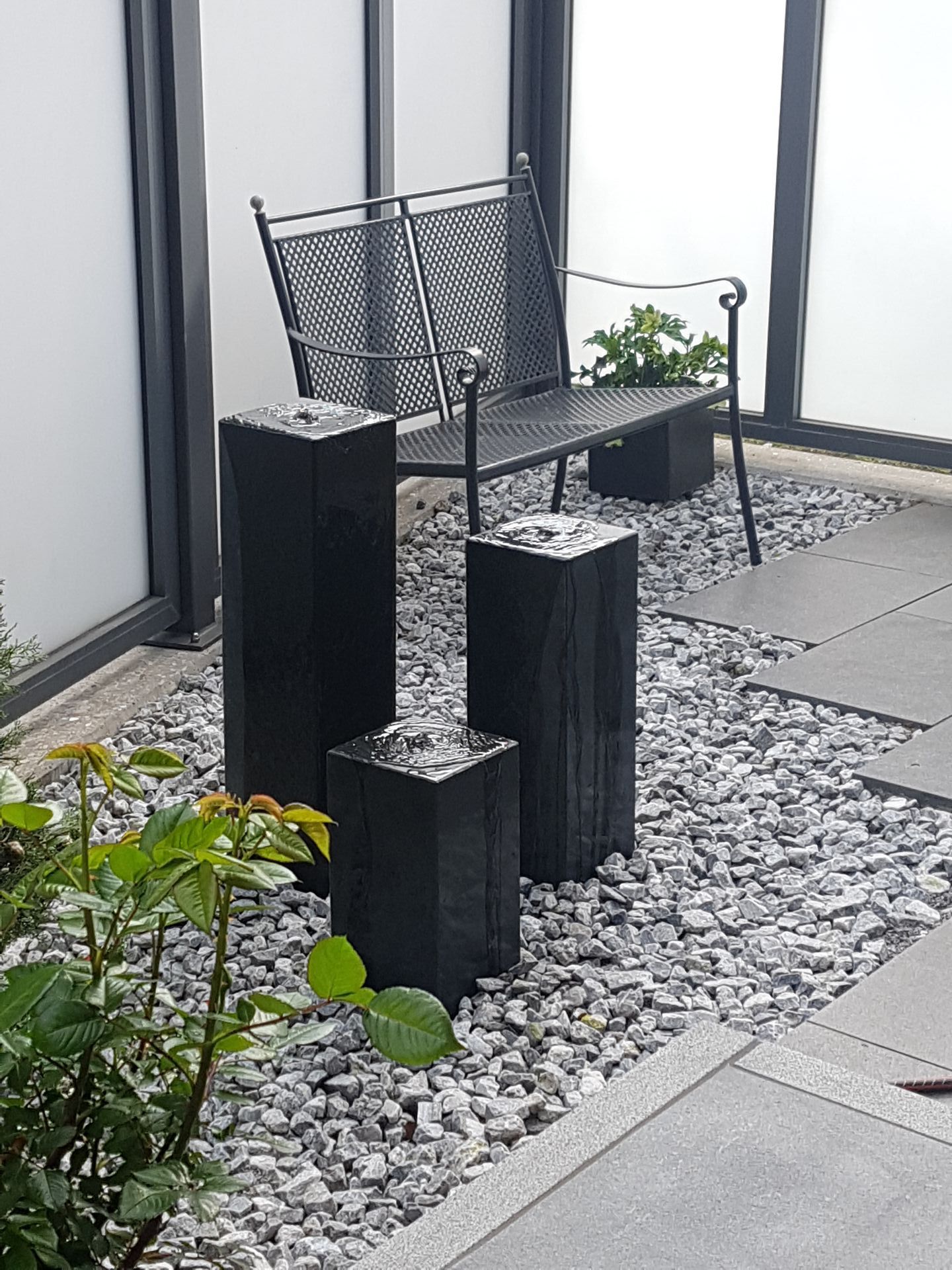Schwarze Wasserfontänen in einem Kiesbett, im Hintergrund eine schwarze Bank.