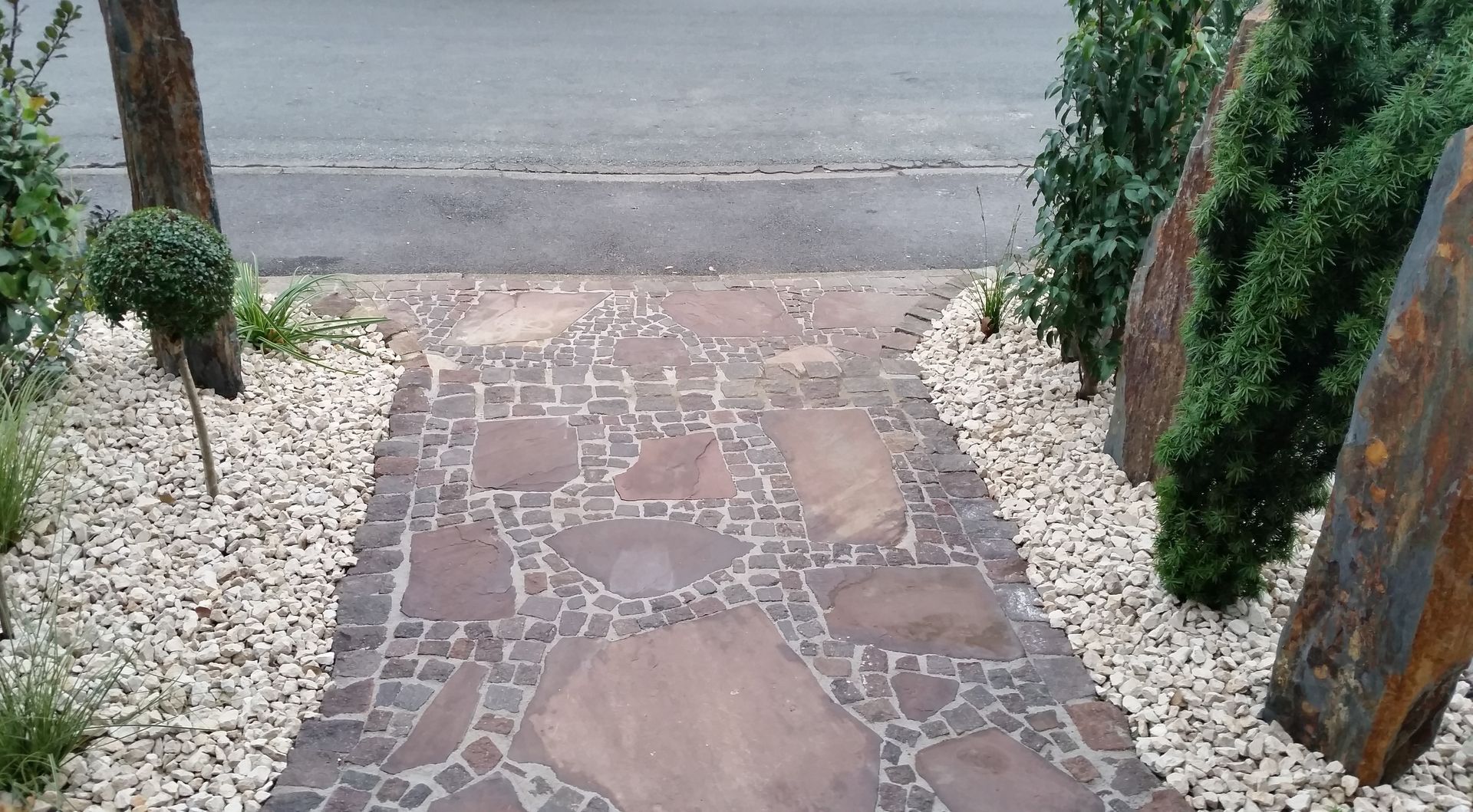 Steinweg, der zu einer Straße führt, gesäumt von weißem Kies und Grün.