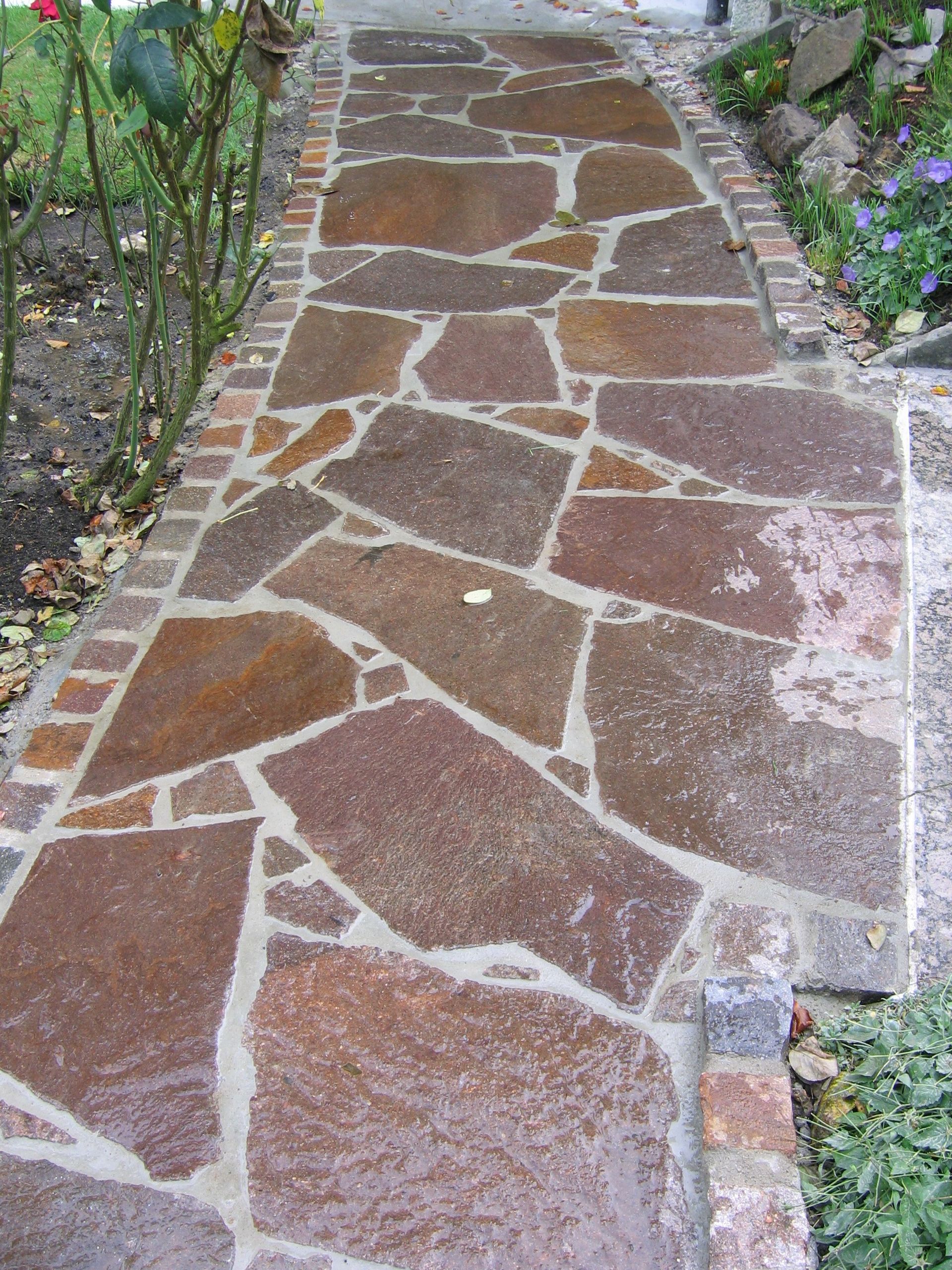 Steinweg mit unregelmäßigen rotbraunen Platten, gesäumt von Ziegeln, der zu einem Gebäude führt.