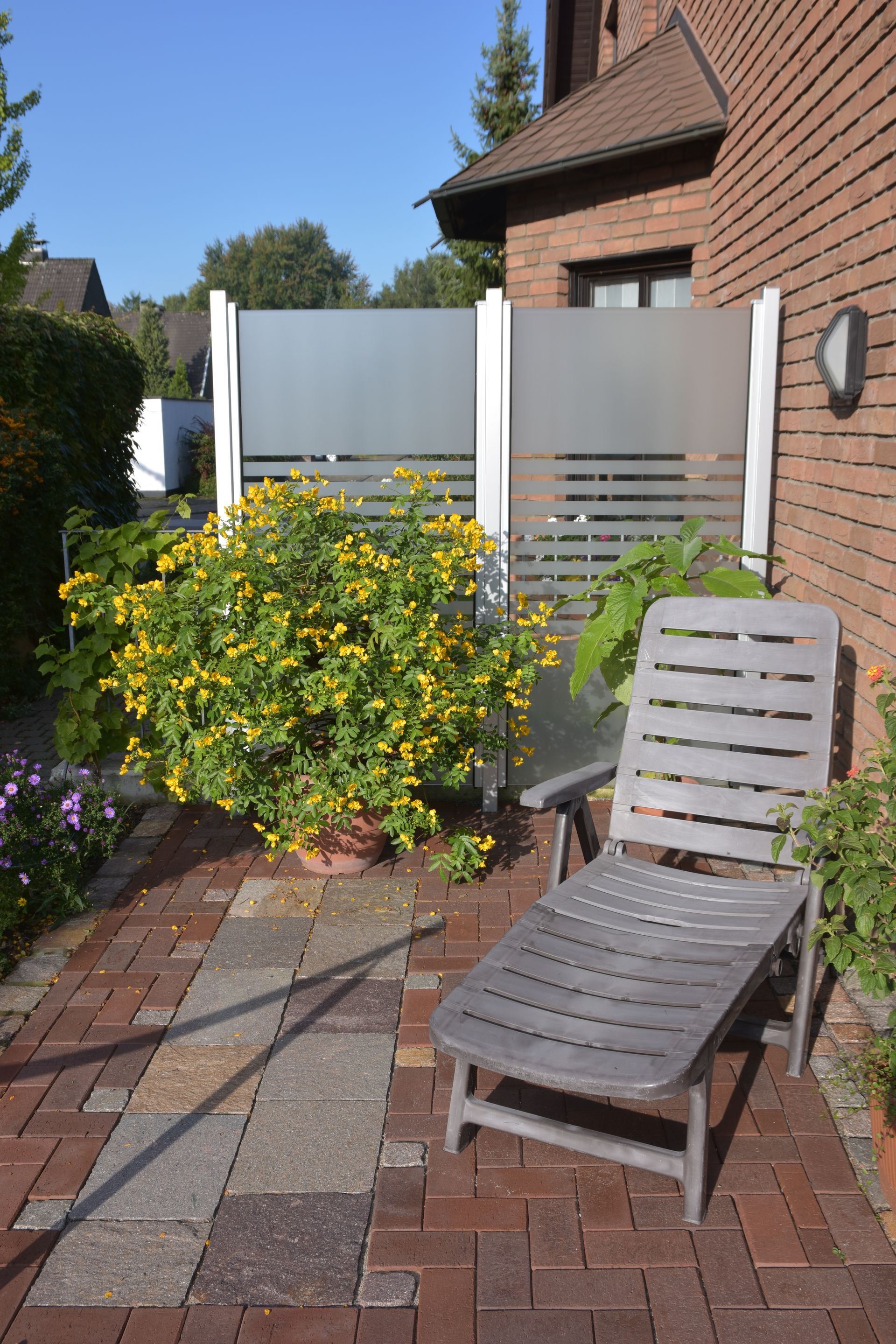 Ziegelterrasse mit Liegestuhl, gelb blühendem Busch und mattiertem Sichtschutz.