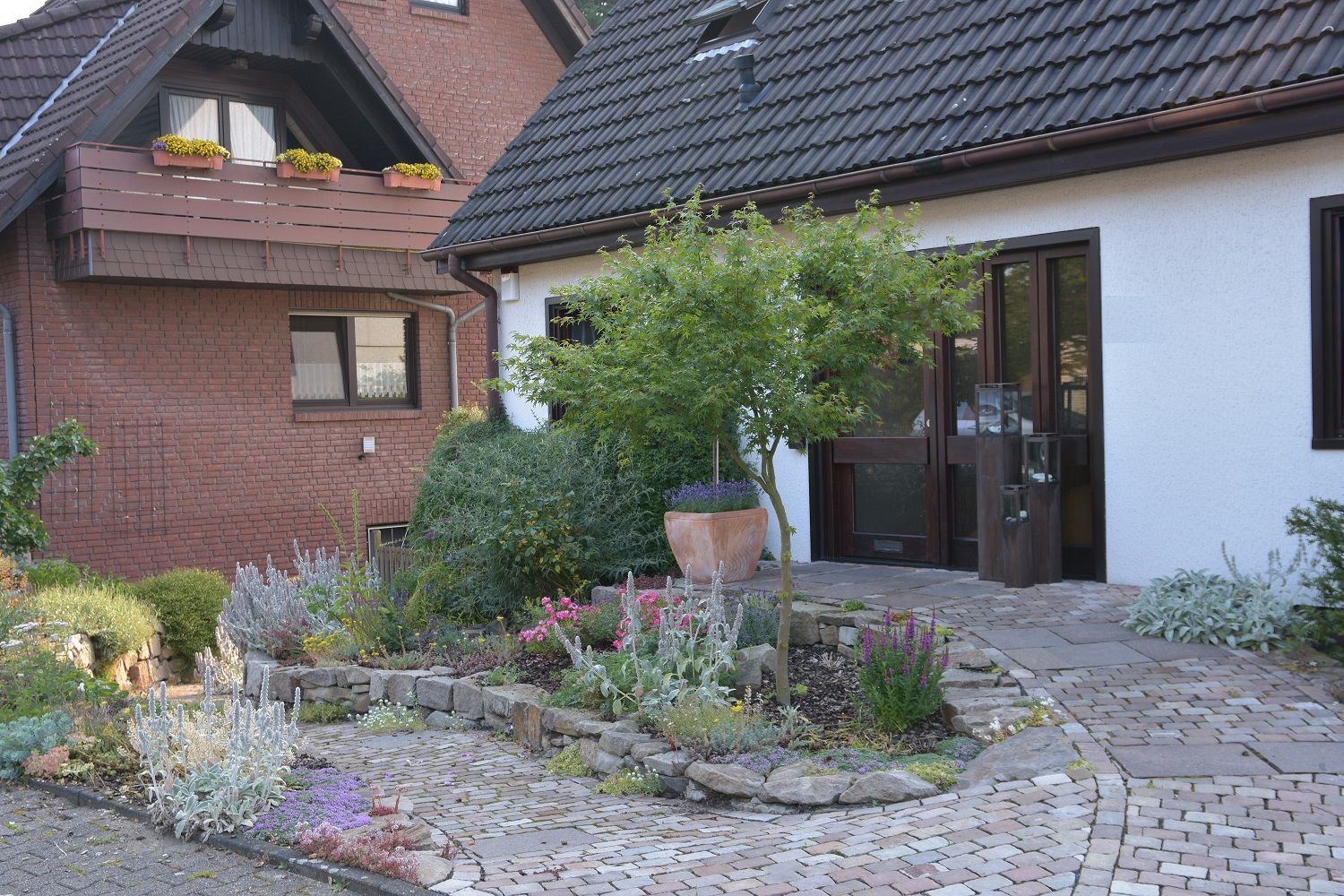 Ein Backsteingebäude mit einem Garten davor. Ein Weg führt zu einem weißen Gebäude mit dunklem Dach.