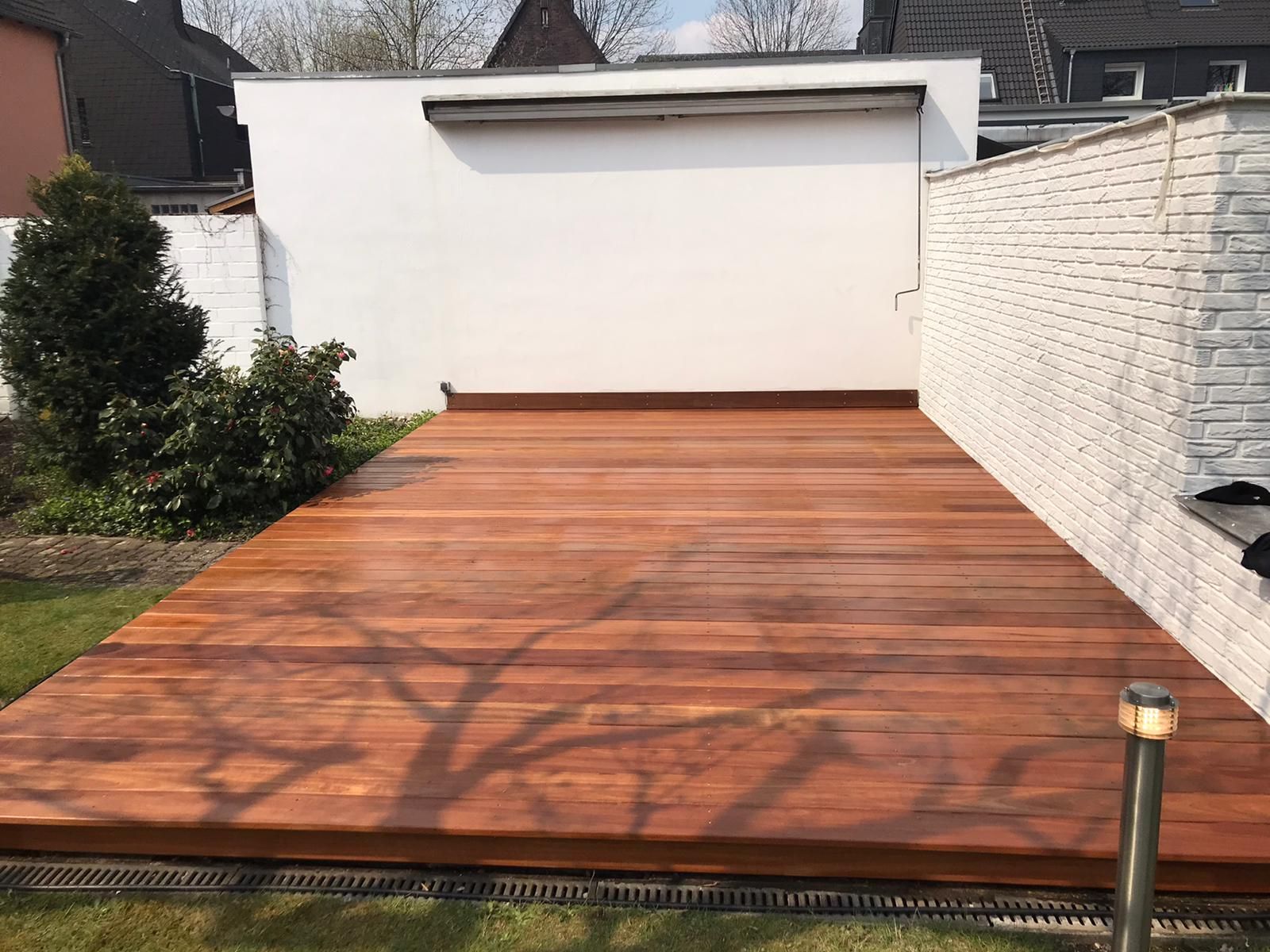 Holzterrasse in einem Hinterhof mit weißer Wand und dekorativer Beleuchtung.