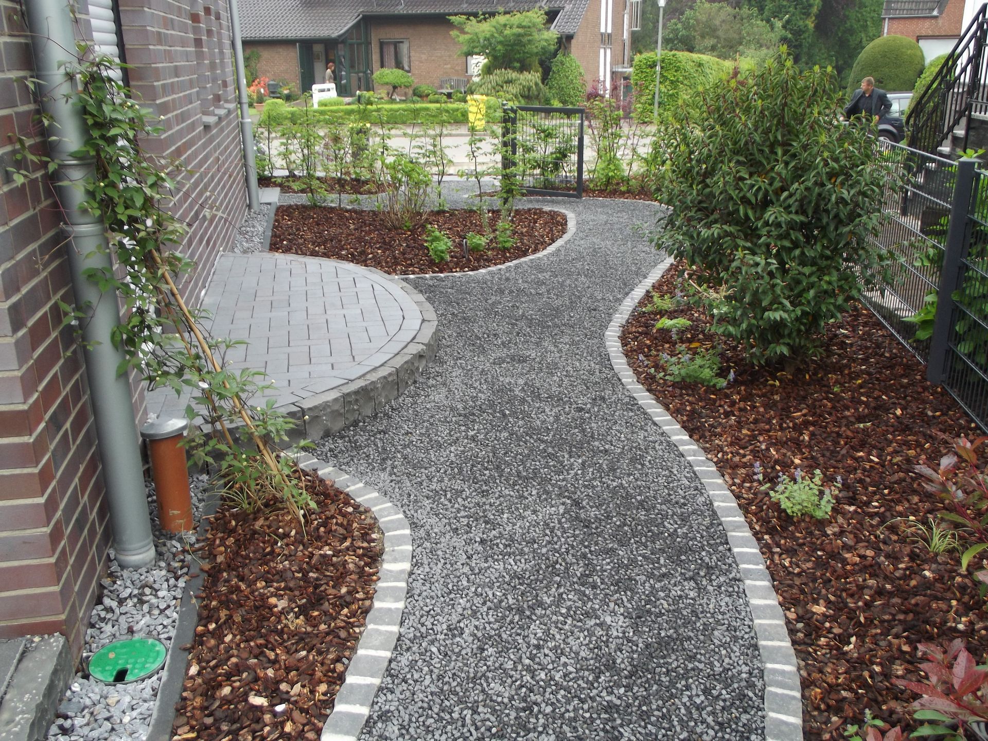 Ein von Pflastersteinen und Mulch gesäumter Kiesweg führt zu einem Backsteingebäude und einem kleinen Garten mit viel Grün.