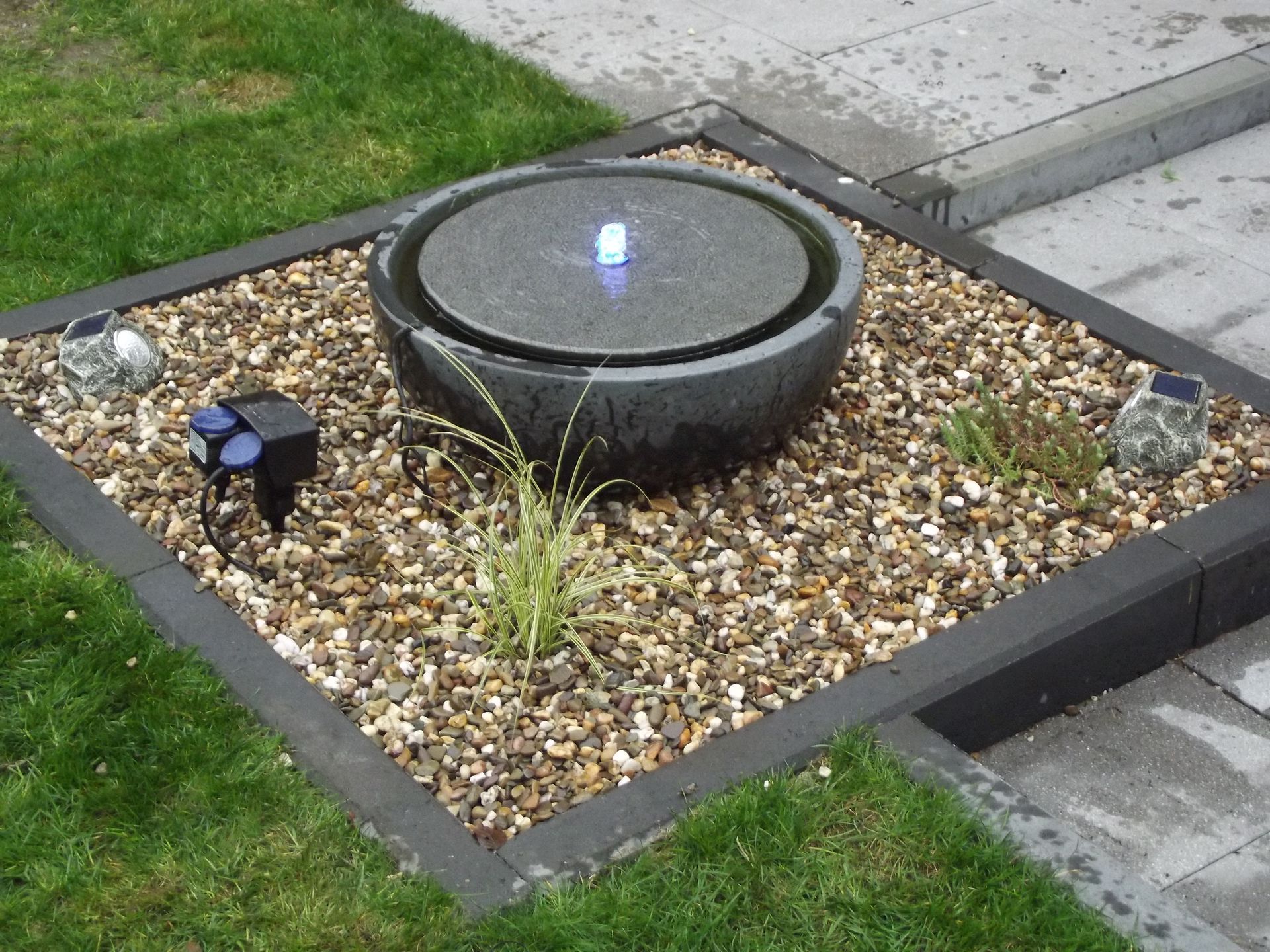 Wasserbrunnen in einer Steinschale, umgeben von Kies und eingerahmt von dunklem Holz, auf einer Rasenfläche.