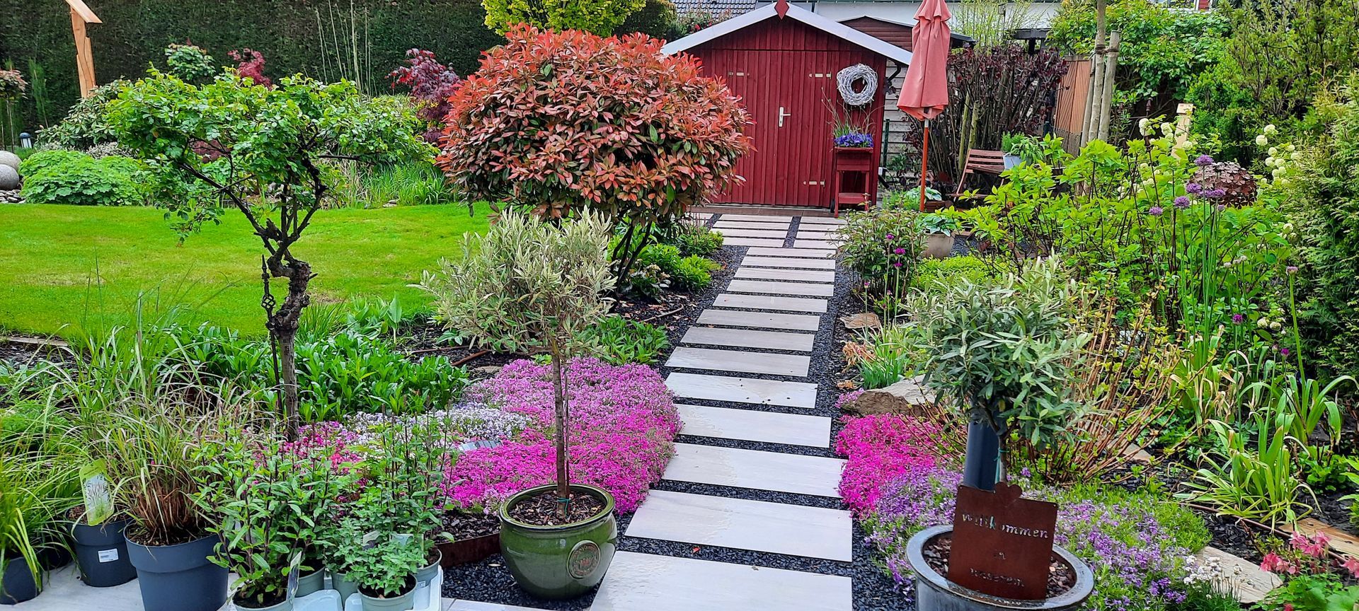 Ein üppiger Garten mit einem Steinweg, der zu einem roten Schuppen führt. Blumen und Grünpflanzen säumen den Weg.