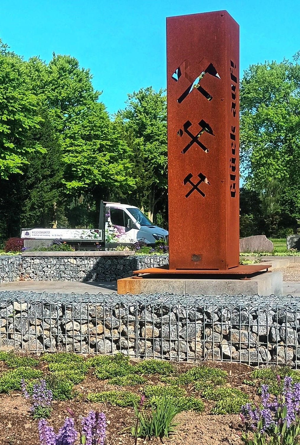 Denkmal aus rostiger Metallsäule mit Bergbausymbolen in einem Park mit einem kleinen Lastwagen im Hintergrund.