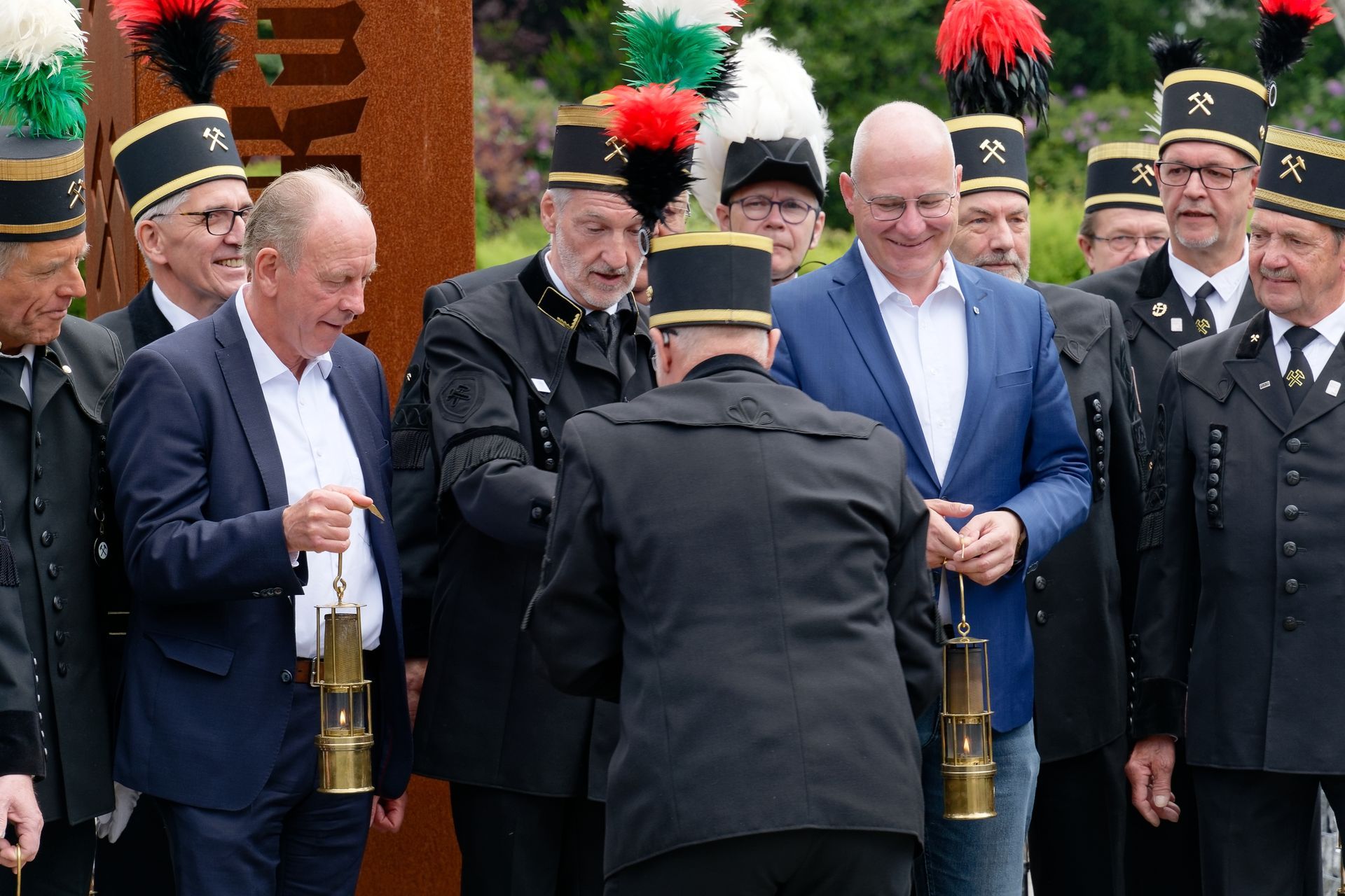 Männer in Bergmannsuniformen und Anzügen versammeln sich draußen mit Laternen an einem Denkmal.