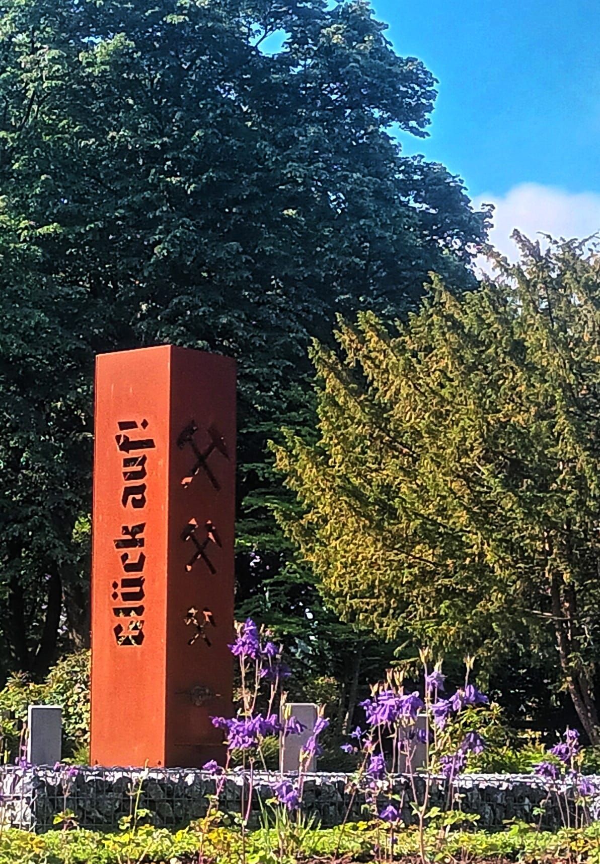 Rostbraunes Denkmal mit dem deutschen Text „Glück auf!“ und Bergbausymbolen, in einem Garten mit lila Blumen und Bäumen.