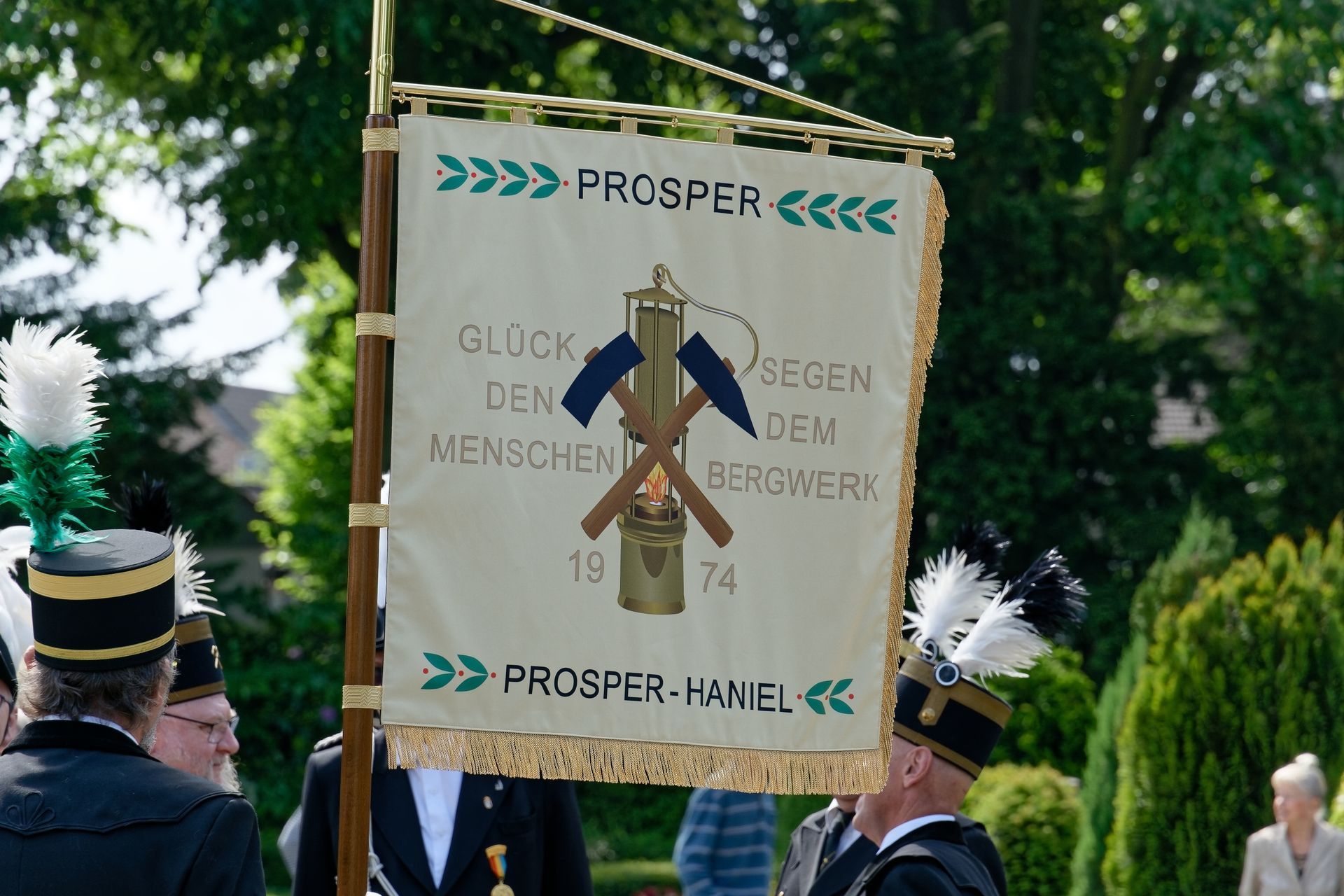 Banner mit „PROSPER“ und gekreuzten Spitzhacken, gehalten von Personen in formeller Kleidung, wahrscheinlich einer Bergbauvereinigung.