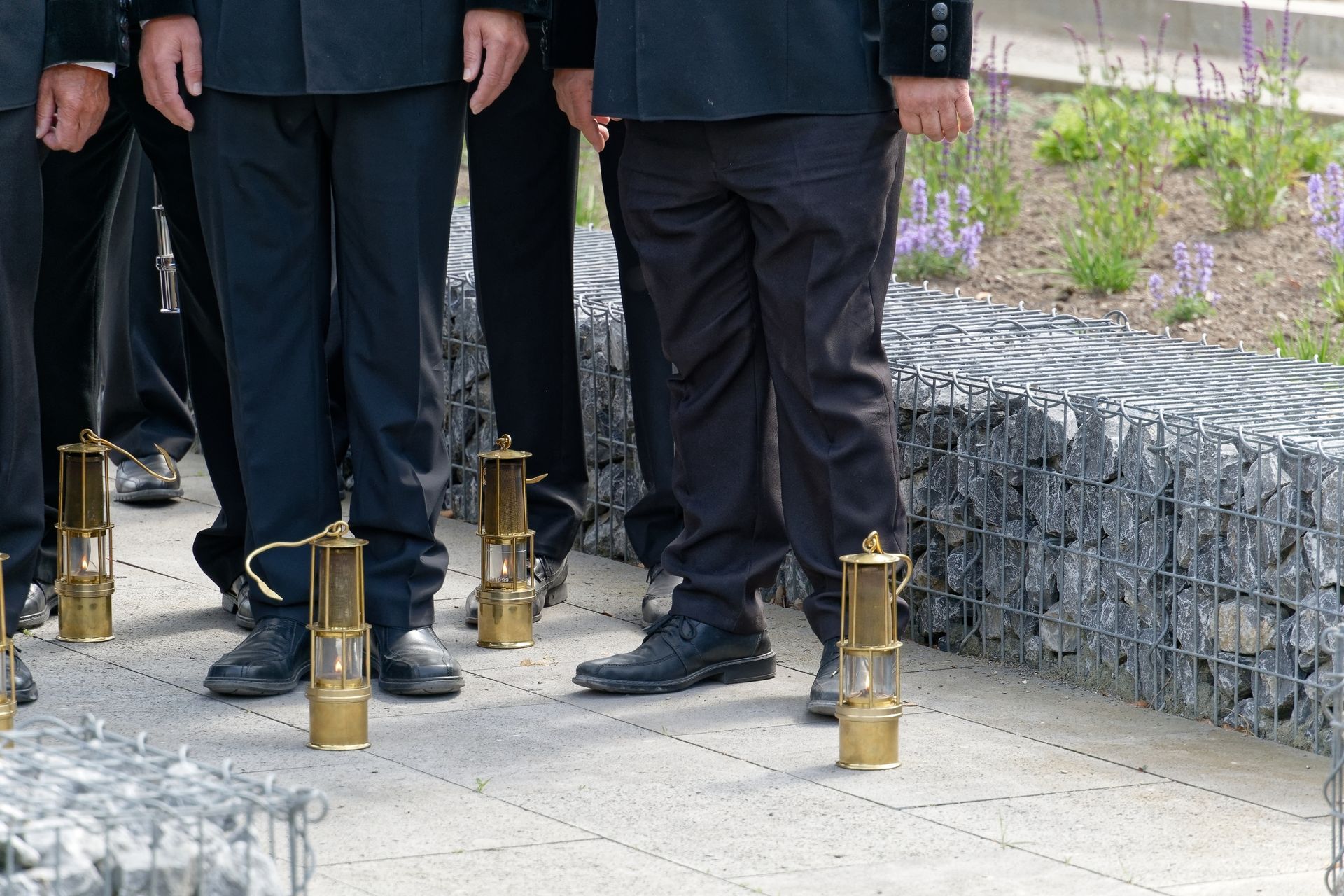 Männer in schwarzen Anzügen stehen mit antiken Grubenlampen auf einem gepflasterten Platz neben einer Felswand.