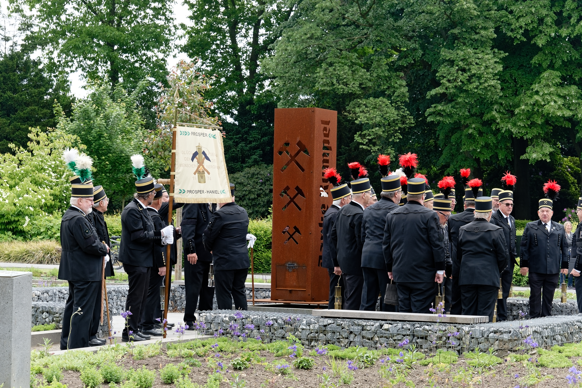 Gedenkzeremonie für Bergleute. Männer in schwarzen Uniformen mit roten Akzenten umgeben im Freien ein Denkmal.