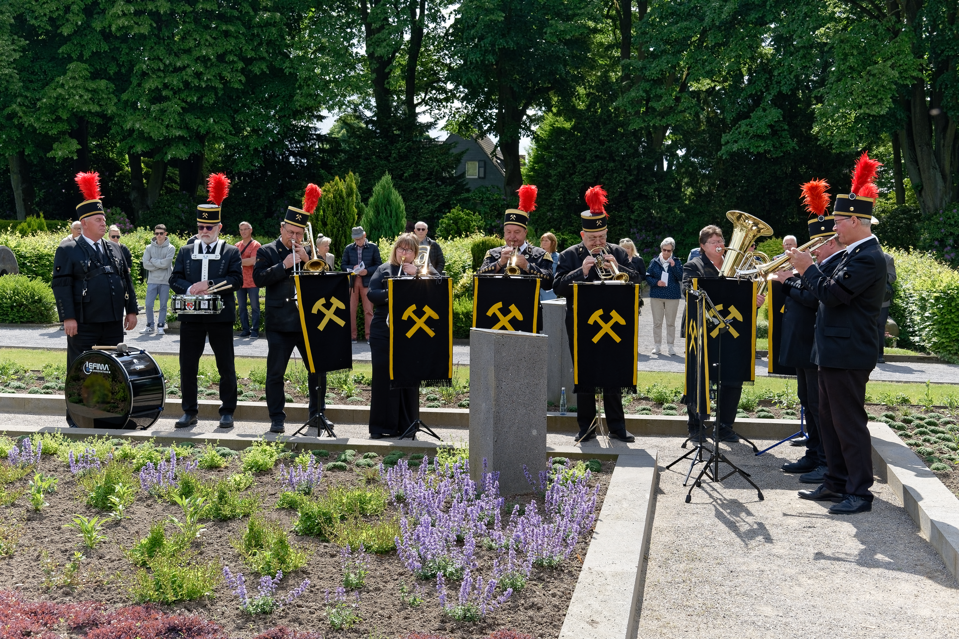 Eine Kapelle in schwarzen Uniformen mit roten Federbüschen und Bergbausymbolen spielt im Freien in der Nähe von Blumen.