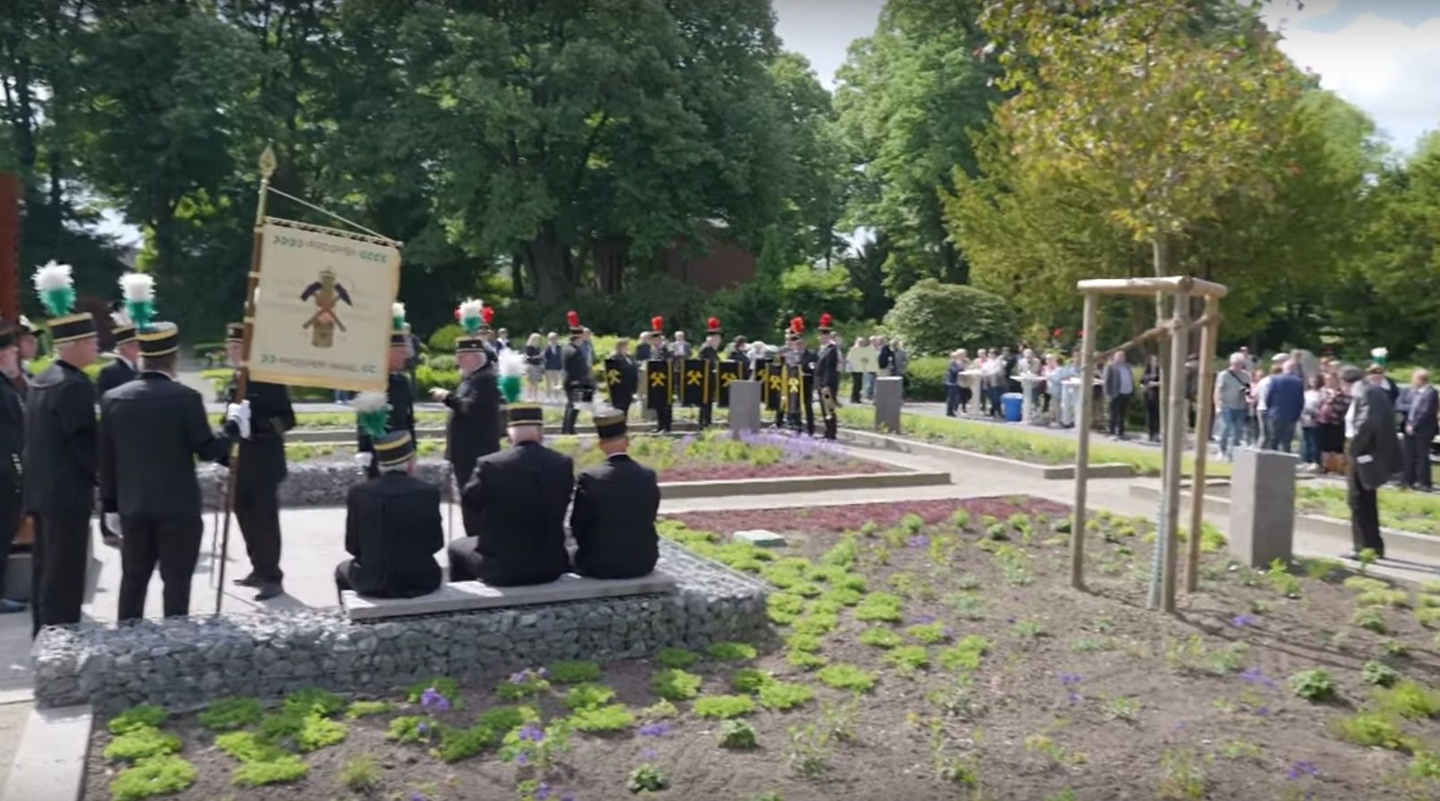 Zeremonie mit einer Band, einer Flagge und Menschen in einem Park. Menschen stehen und sitzen, einige in Uniform.
