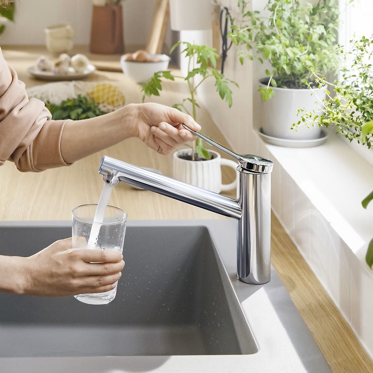 Une personne remplit un verre sous un robinet de cuisine, à côté d'un évier, avec des herbes en pot près de la fenêtre.