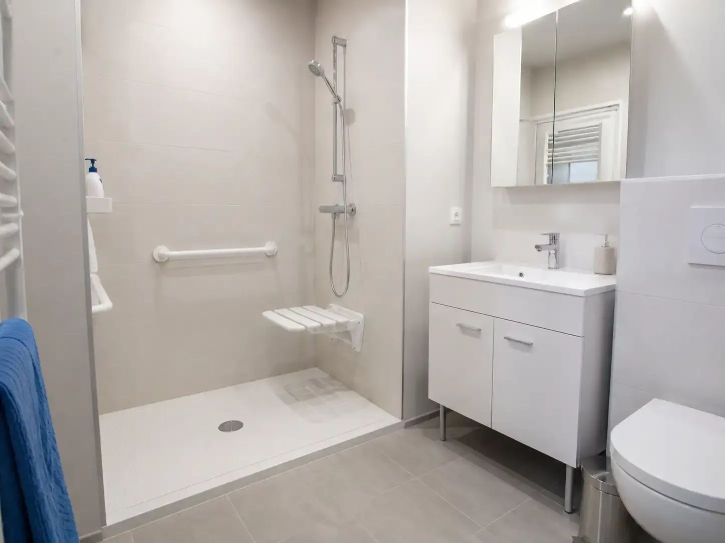 Salle de bain moderne et accessible avec douche à l'italienne, lavabo, miroir et toilettes dans des tons blancs et gris.