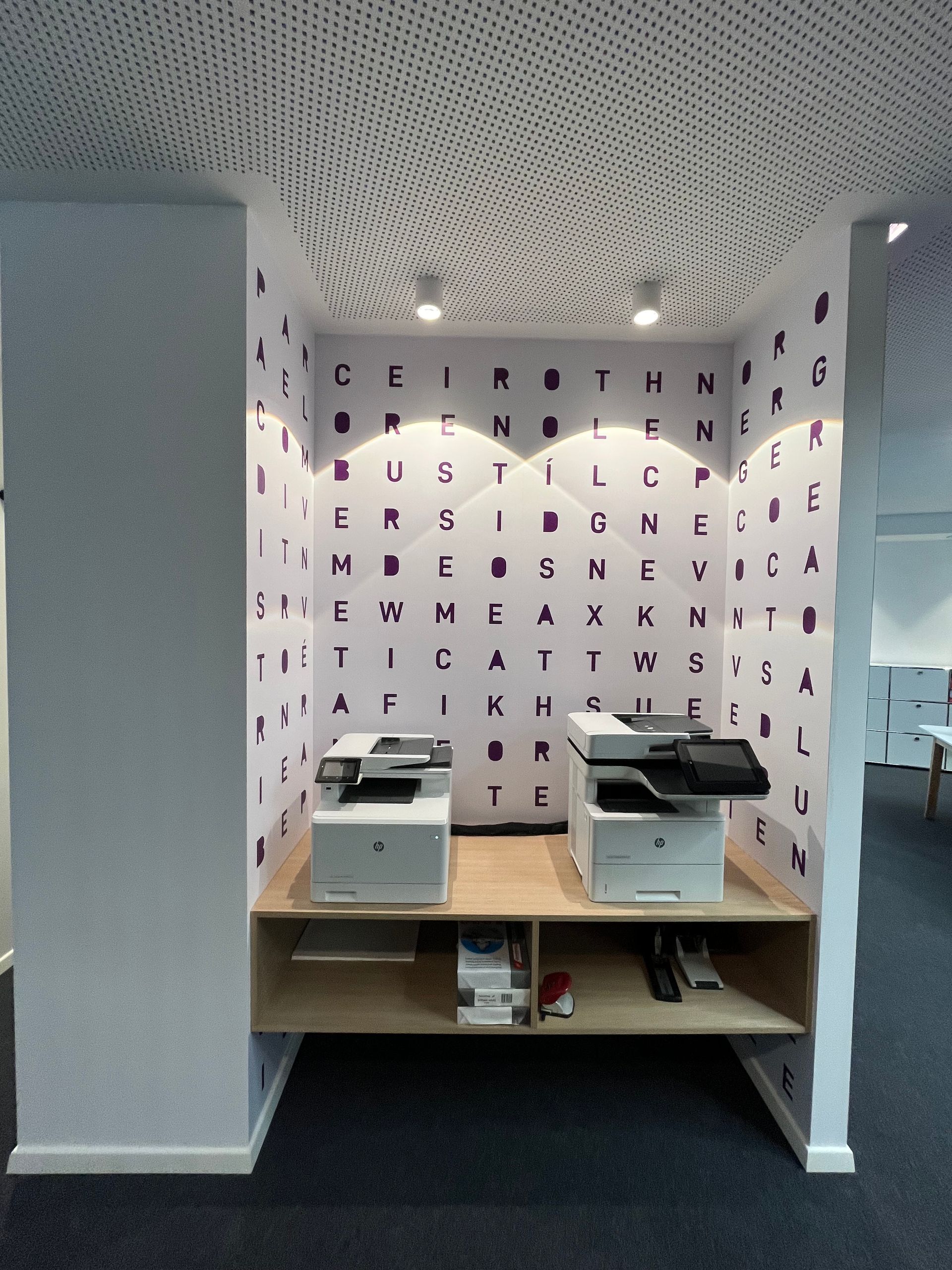Two printers are on a shelf in front of a wall with letters on it