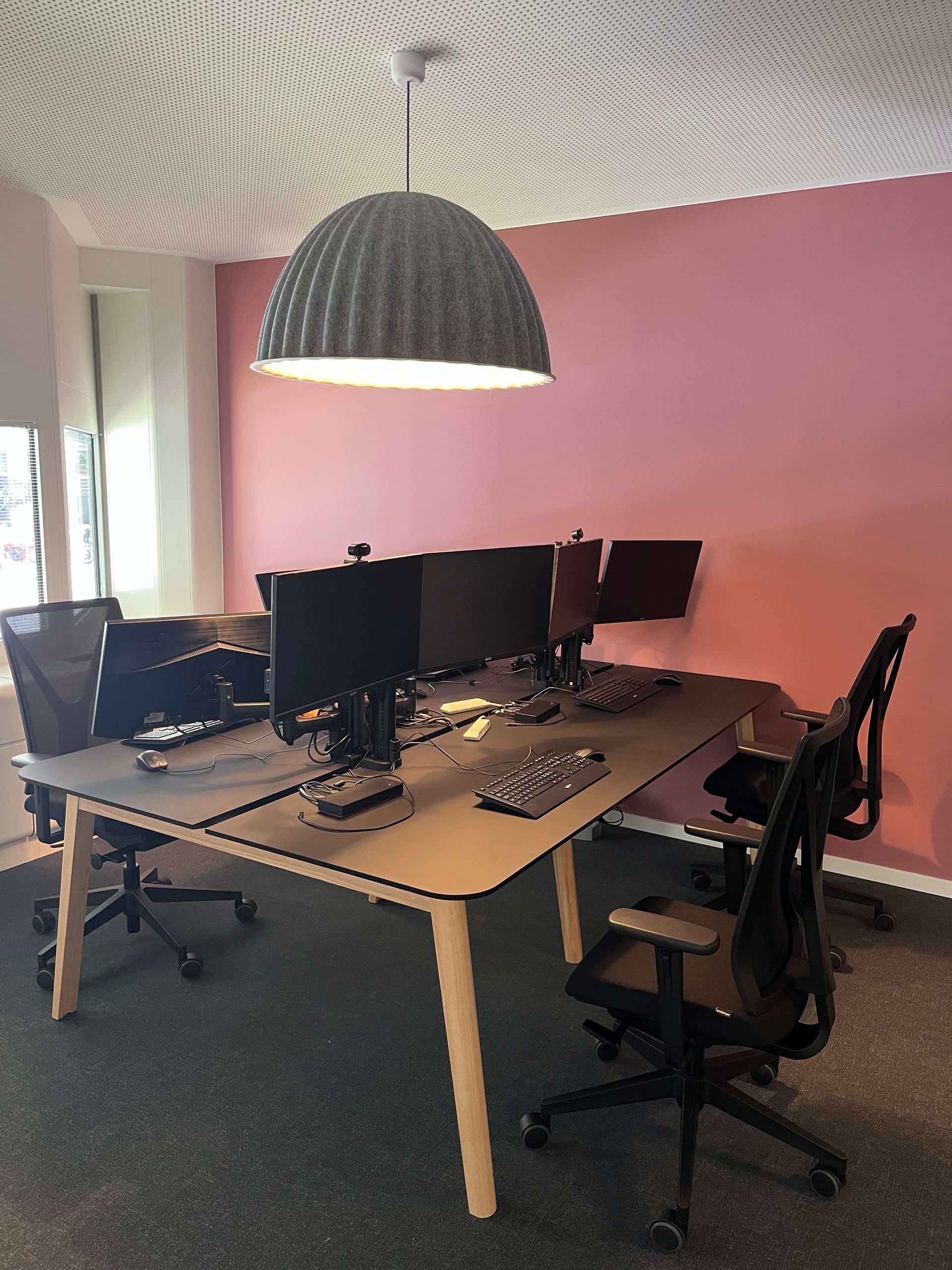 A row of desks with computers and chairs in a room with a pink wall.