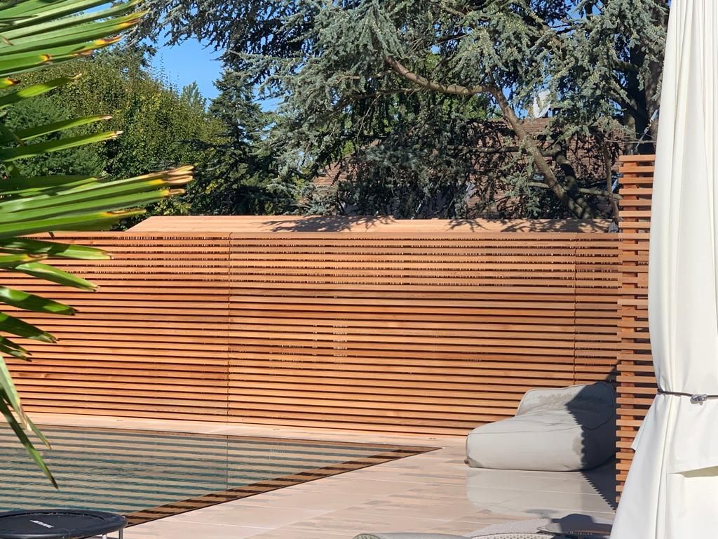 A wooden fence surrounds a swimming pool with a couch and umbrella in front of it.