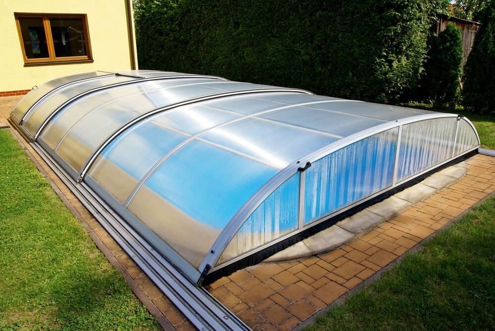 Piscine extérieure couverte, toit arqué translucide, pelouse verte, bordure en briques et maison en arrière-plan.