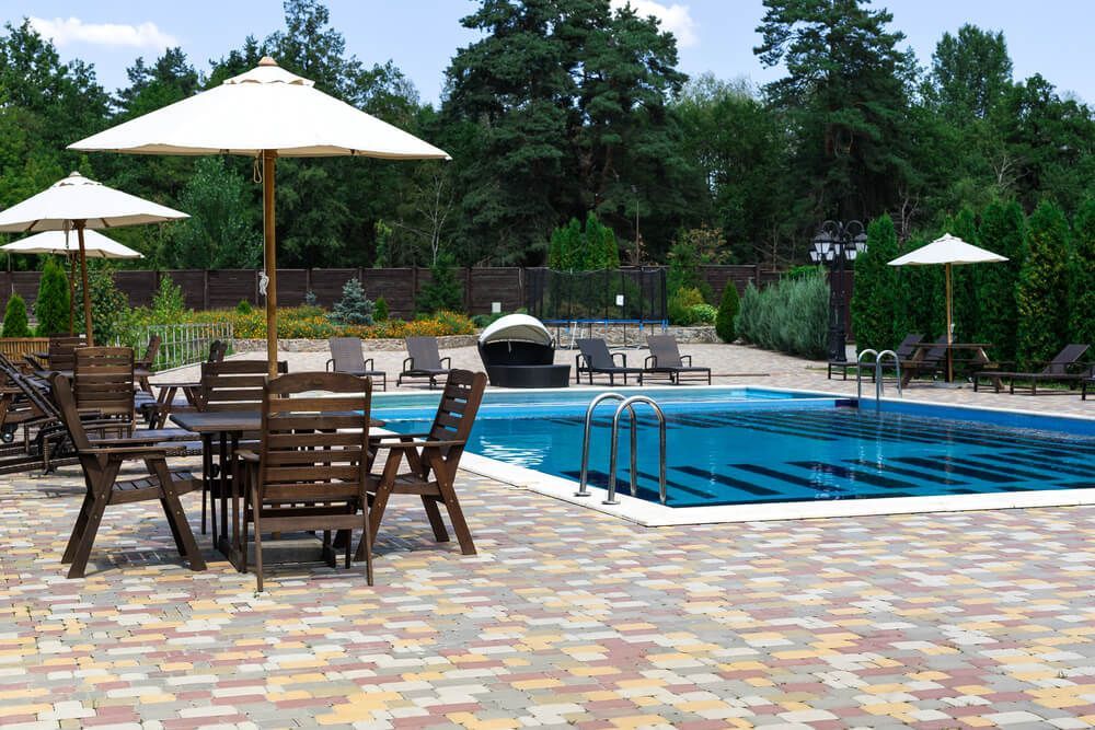 Scène au bord de la piscine avec tables, chaises, parasols et transats près d'une piscine bleue, entourée d'arbres.