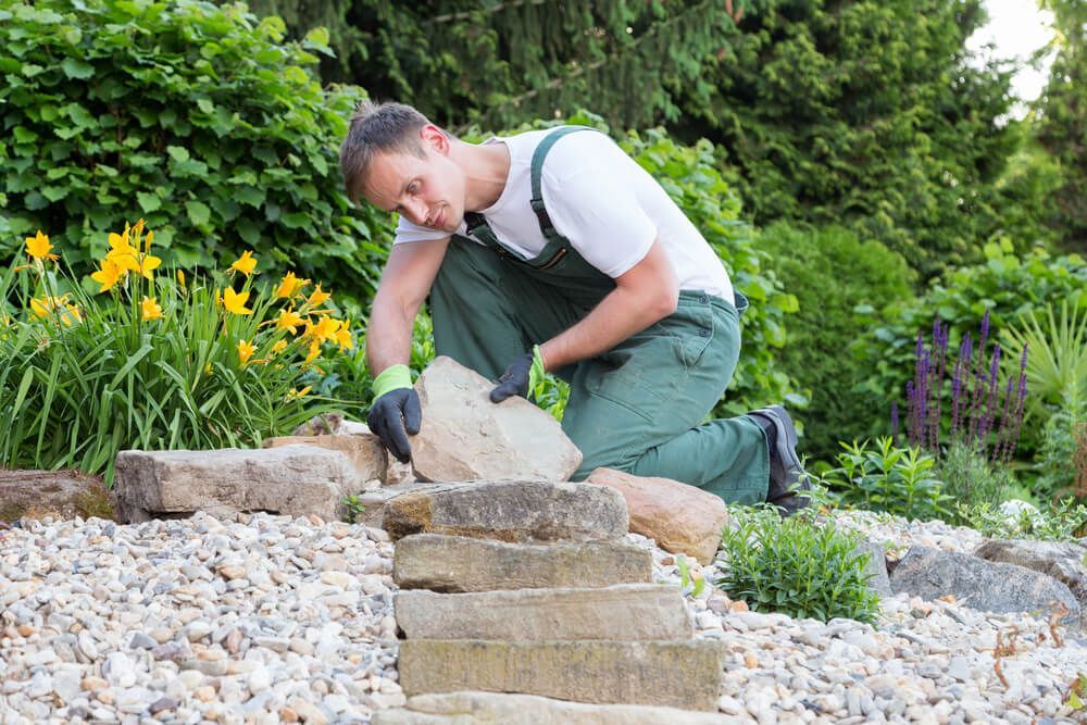 Un homme en salopette verte dépose une pierre dans un jardin fleuri et gravillonné.