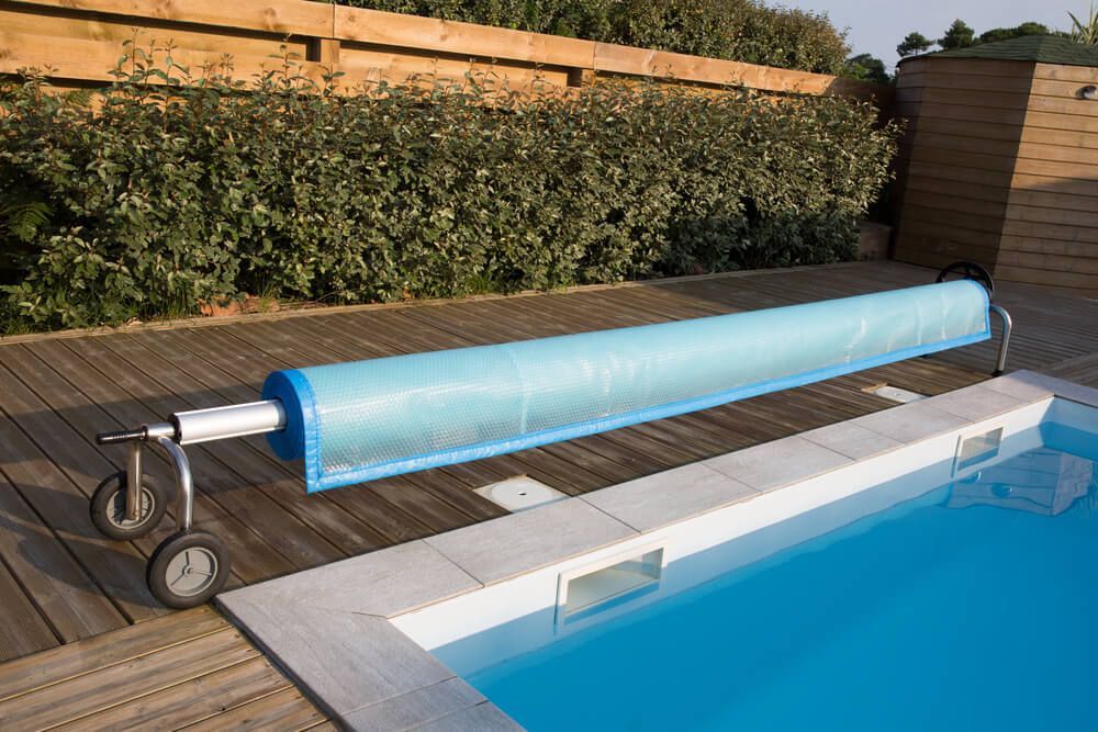 Piscine avec une bâche bleue enroulée sur une barre métallique, à côté d'une terrasse en bois et d'arbustes.