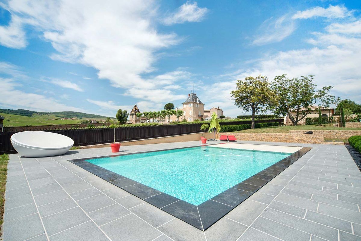 Piscine à l'eau bleue et terrasse en pierre, avec un château en arrière-plan sous un ciel bleu.