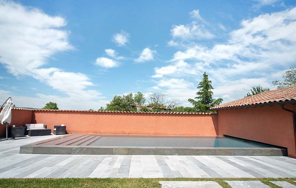 Une piscine rectangulaire entourée d'une terrasse en pierre grise, adossée à un mur orange sous un ciel bleu.