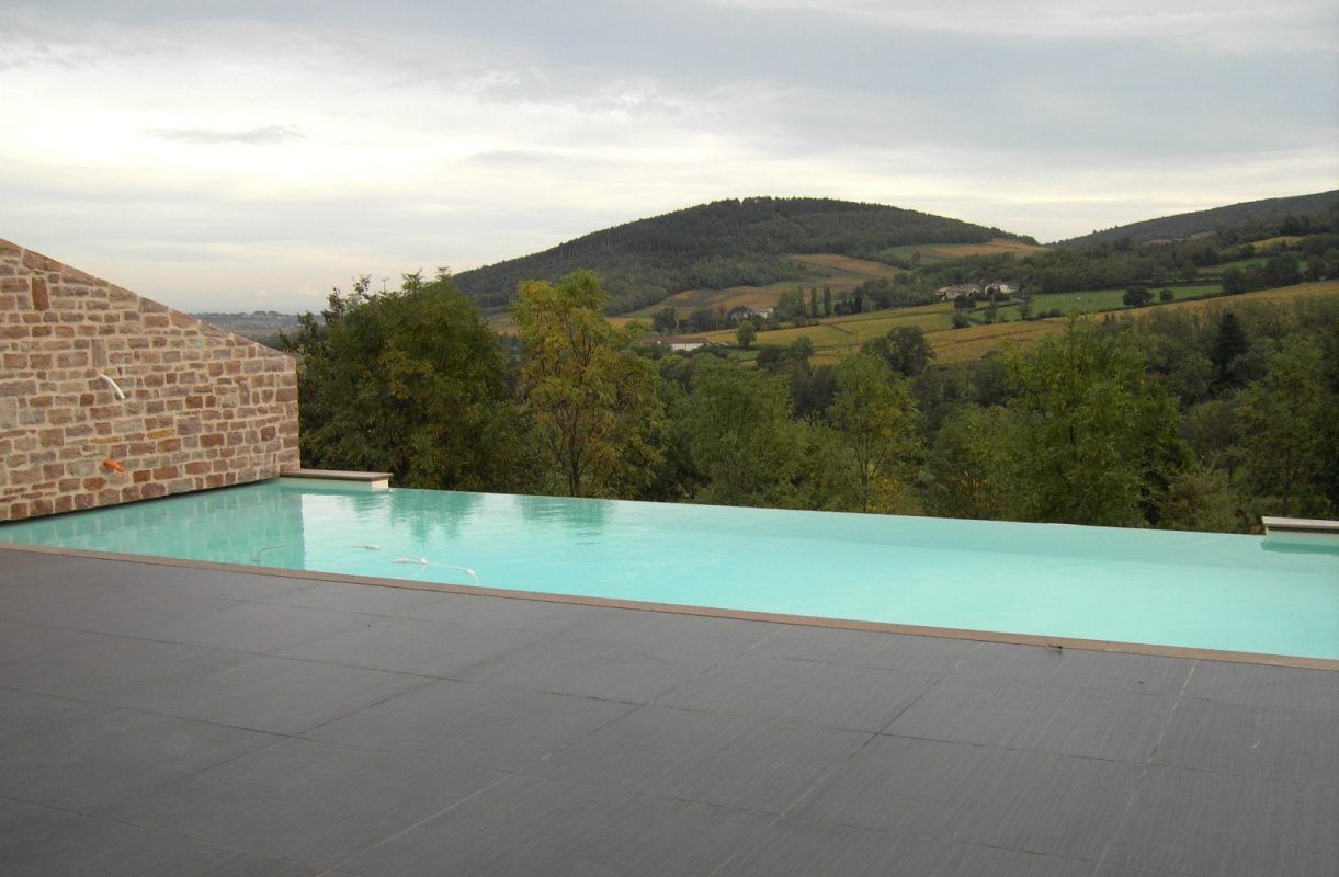 Piscine à débordement donnant sur des collines verdoyantes et des arbres. Mur de pierre à gauche, ciel couvert.