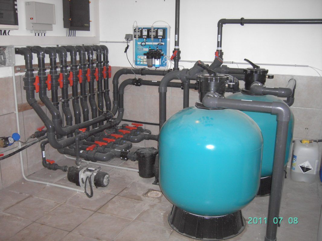 Système de filtration de piscine avec réservoirs bleus, tuyaux noirs et vannes orange dans un local technique.