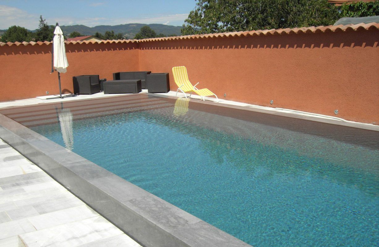 Piscine extérieure rectangulaire à l'eau turquoise, entourée d'une terrasse grise et d'un mur rouge.