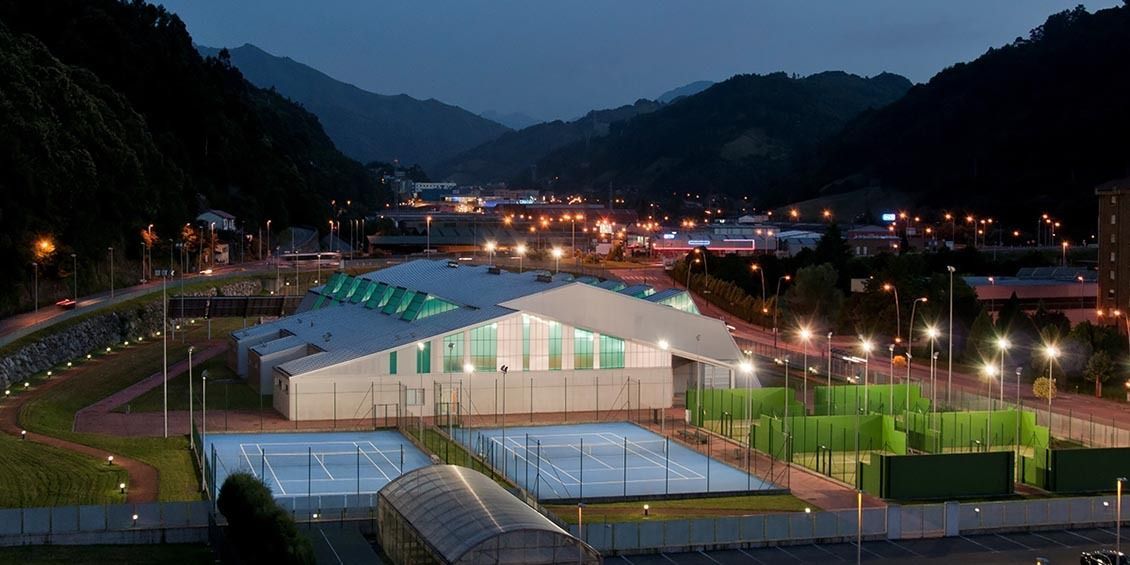 Complejo deportivo iluminado con edificios de techos verdes en un valle de montaña al atardecer.