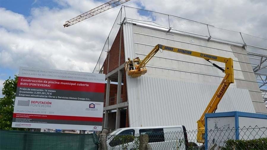 Obra en construcción con grúa, plataforma elevadora amarilla y edificio blanco junto a un letrero rojo bajo un cielo nublado.