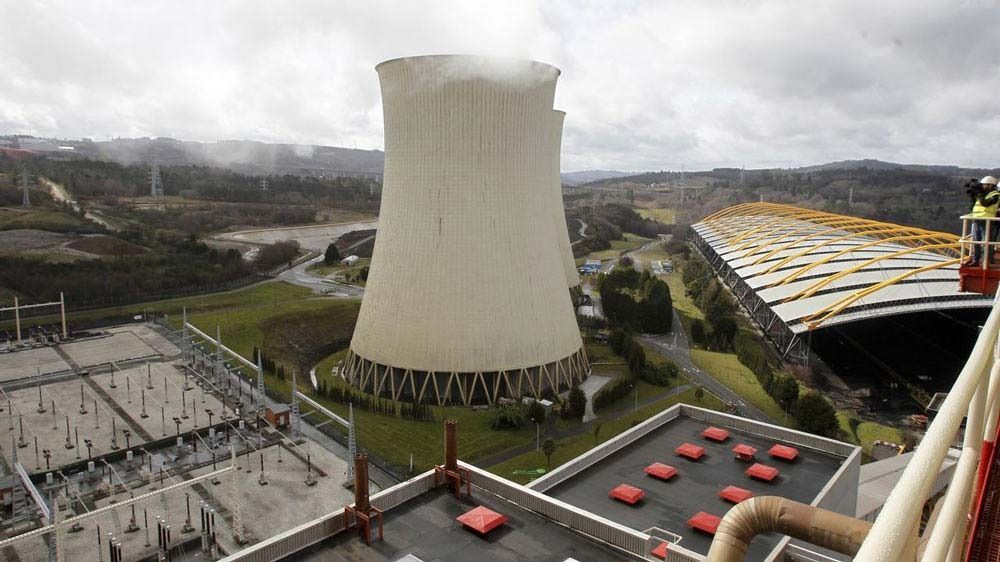 Central eléctrica con una gran torre de refrigeración, edificios industriales y trabajadores en una ladera bajo un cielo nublado.