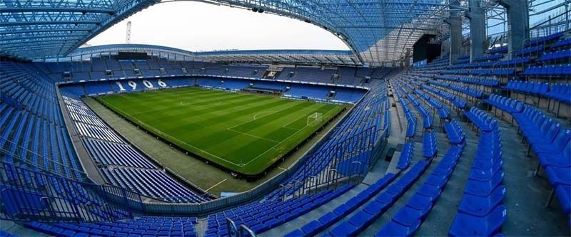Estadio de fútbol vacío con asientos azules y un campo verde bajo un techo curvo.