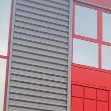 Fachada de edificio roja y gris con ventanas y un panel de celosía vertical.