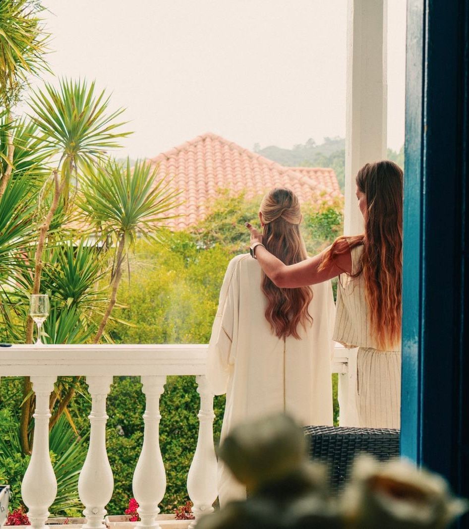 Dos mujeres están paradas en un balcón mirando hacia un exuberante campo verde.