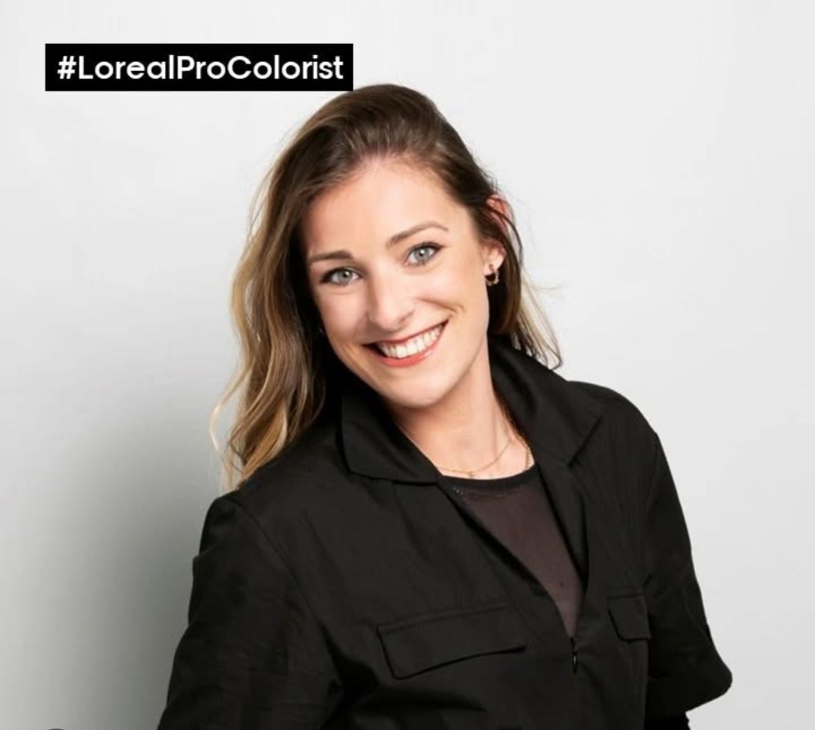 Una mujer que lleva una chaqueta negra sonríe con un logotipo #lorealprocolorist detrás de ella.