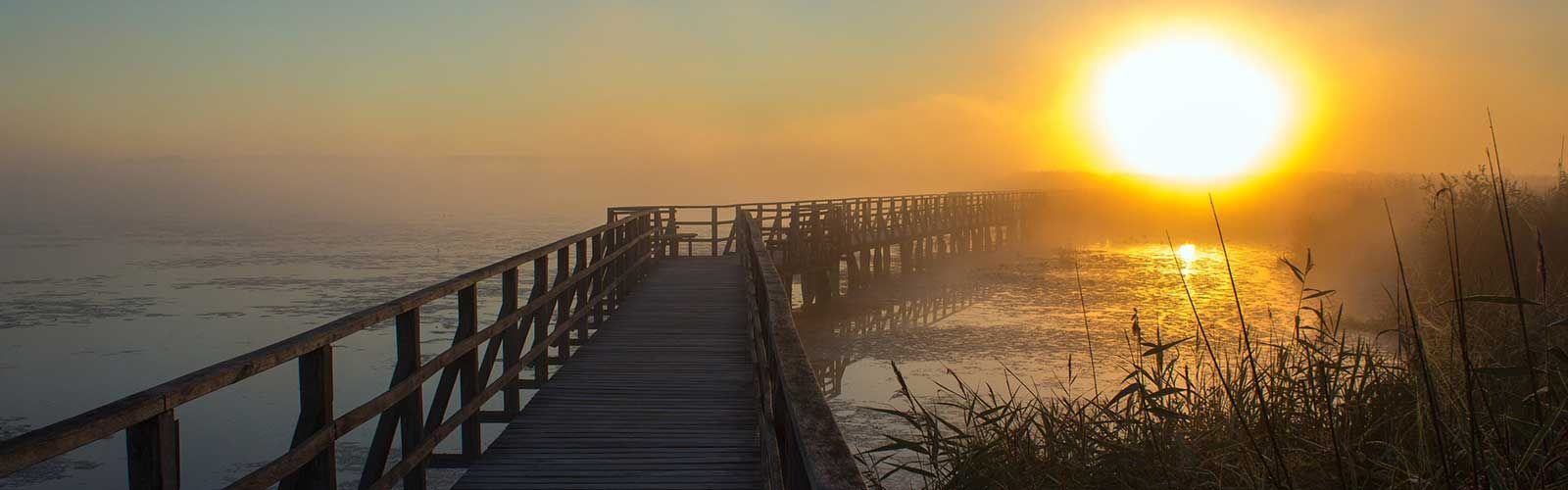 Die Sonne geht über einem See unter, im Vordergrund ist ein Holzsteg zu sehen.