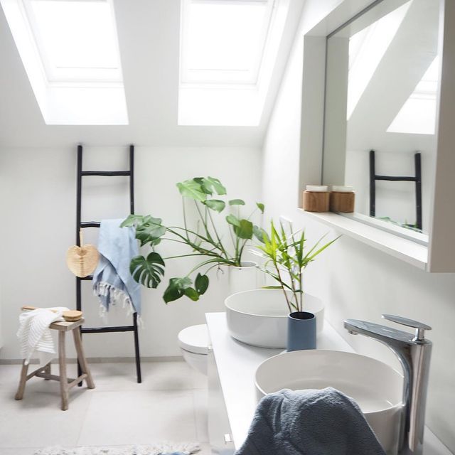 Baño blanco brillante con tragaluces, lavabo, plantas y escalera negra para toallas.