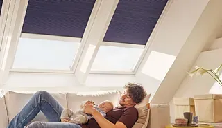 Un hombre sostiene a un bebé, relajándose en un sofá debajo de tragaluces con cortinas azules en una habitación del ático.