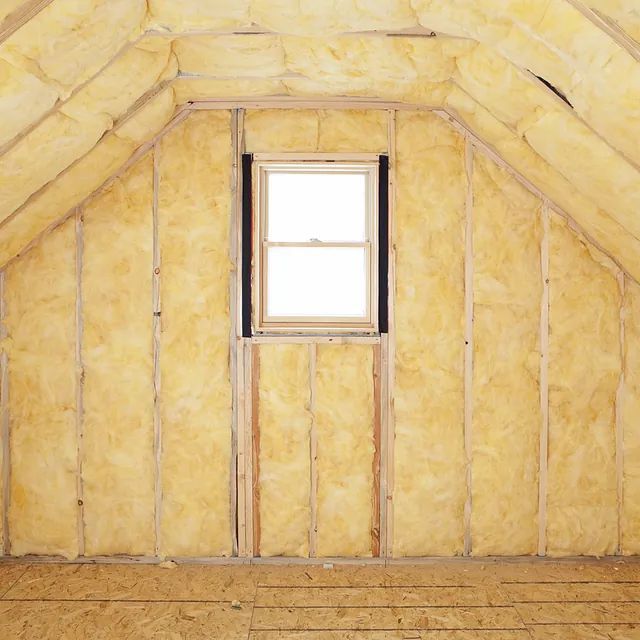 Interior de ático con aislamiento amarillo entre vigas de madera. Pequeña ventana centrada en la pared.