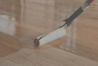 Una persona que utiliza un rodillo de pintura para aplicar una capa transparente a un piso de madera.