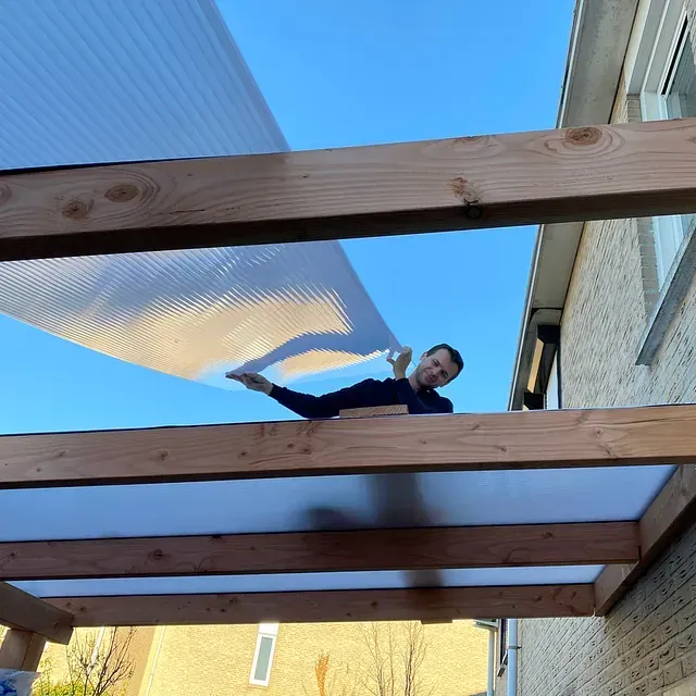 Hombre instalando paneles de techo de plástico corrugado en una pérgola de madera, contra un cielo azul.