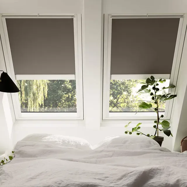 Dormitorio con dos tragaluces, persianas grises, cama blanca y vegetación.