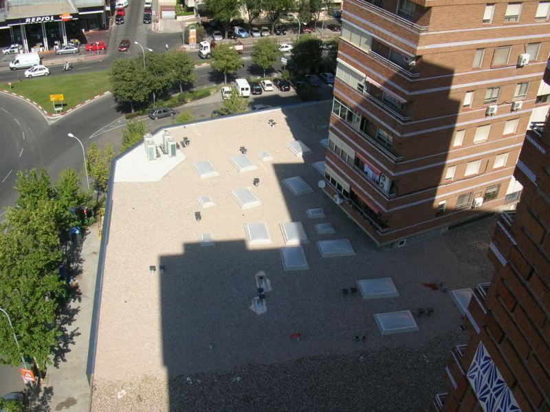 Vista aérea de un edificio con techo plano y tragaluces, junto a un edificio de ladrillo más alto y una calle.