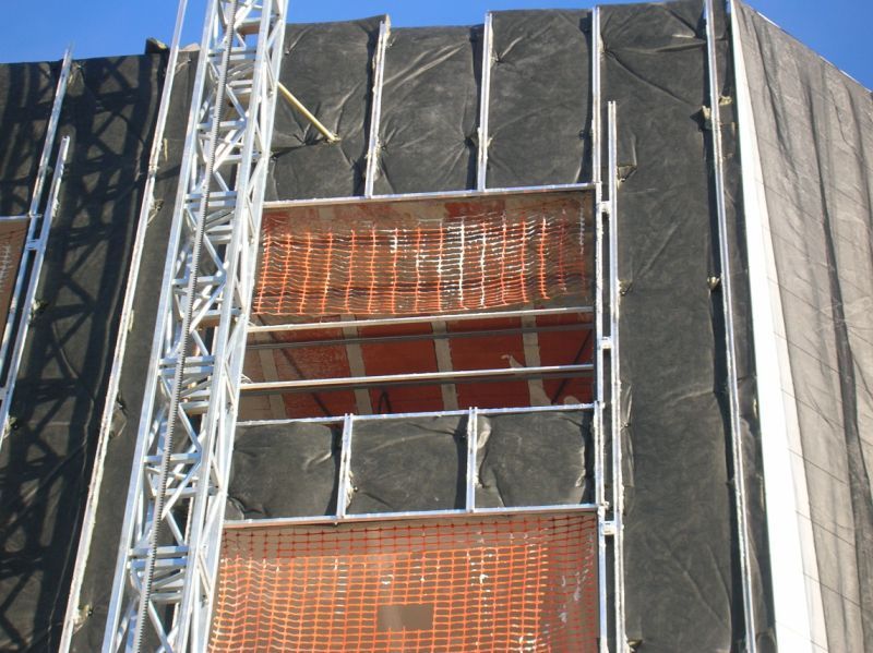 Andamios de construcción en un edificio con ladrillo y revestimiento negro; cielo azul en el fondo.