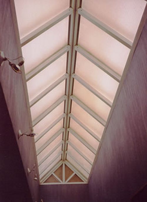 Mirando hacia arriba a un tragaluz con vigas blancas y paneles translúcidos en un pasillo con paredes texturizadas de color púrpura.